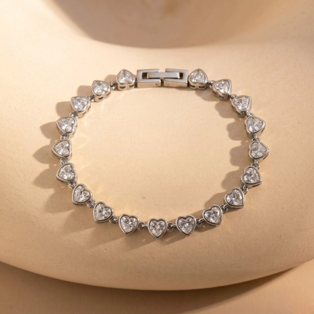 RITI silver tennis bracelet featuring heart-cut cubic zirconia stones in a bezel setting on a beige background.