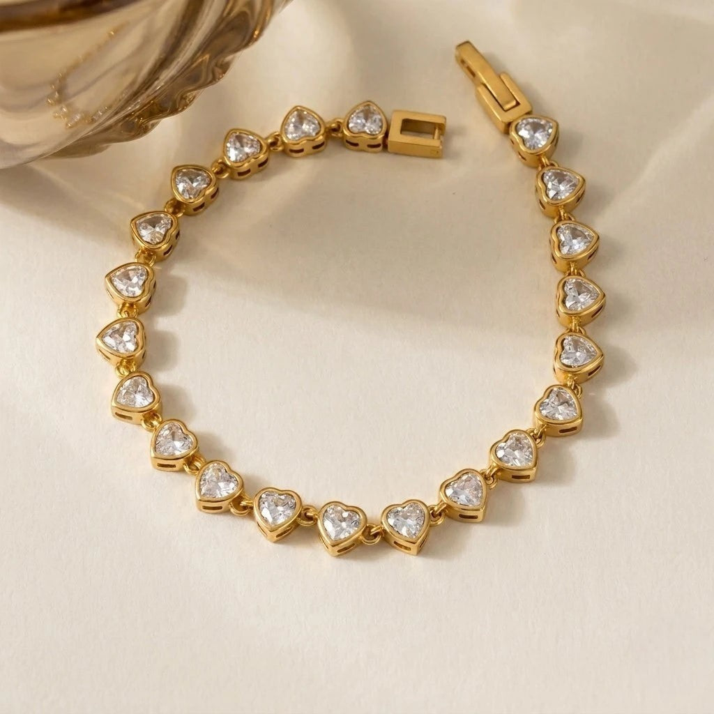 RITI gold tennis bracelet featuring heart-cut cubic zirconia stones in a bezel setting on a soft cream background.
